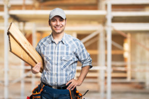 young and happy construction contractor holding planks of wood standing in front of a building remodeling project