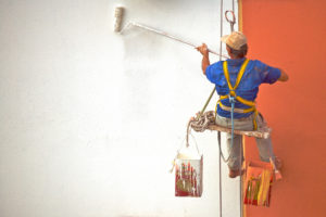 Professional painting contractor painting walls with white and orange as he sits on a swing-like seat to keep him in position to paint.